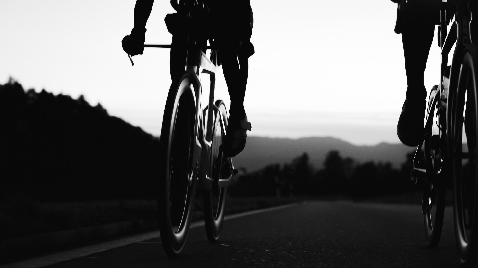 Two road cyclists silhouetted against a bright sky at dawn, shot low from behind on an empty road, in black and white.